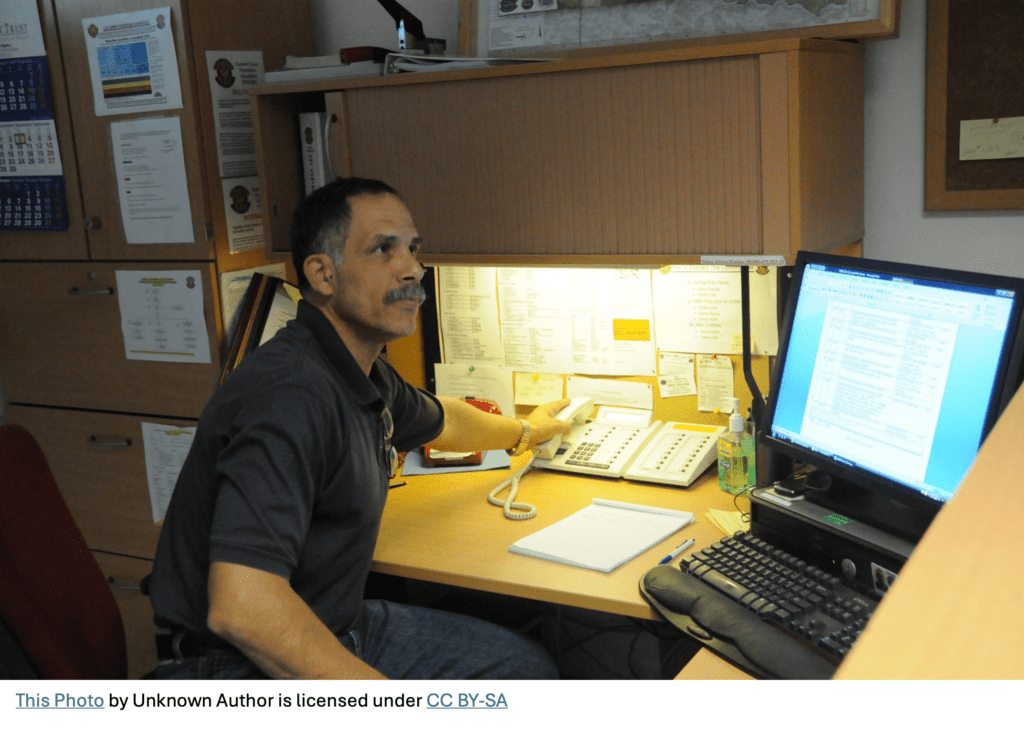 A photo of a project manager at his desk hanging up a phone call and working on a project phase gate document on his computer.